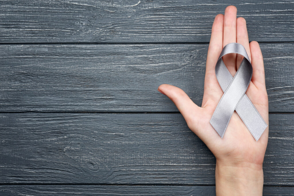 Female hand holding grey ribbon on wooden background. Brain Cancer